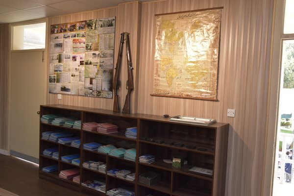 Shelves of bound registers beneath a service rifle and a large framed document mounted on the wall
